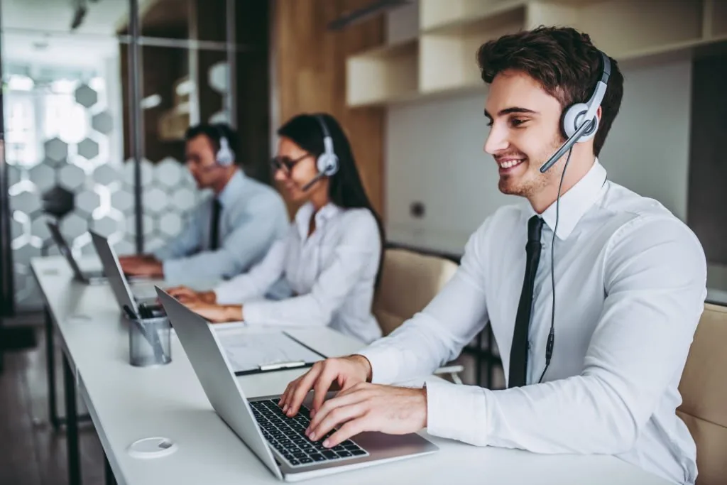 A smiling call center agent in a white shirt and tie typing on a laptop with his headset on, colleagues on calls behind him.